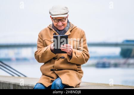 Outdoor portrait of cheerful senior man in winter coat who is using digital tablet. Stock Photo