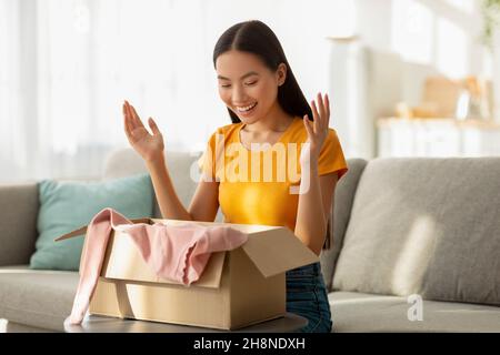 Unpacking clothes. Portrait of happy asian lady opening cardboard box, taking out new shirt, sitting on sofa at home Stock Photo