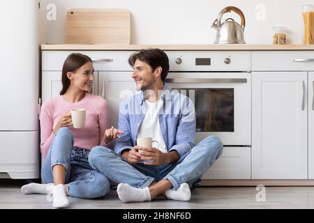 Smiling glad millennial caucasian female and man sit on floor with cups of hot drink talking in modern kitchen Stock Photo