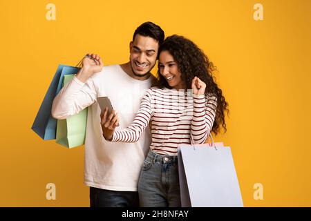 Cashback. Joyful Arab Couple Holding Shopping Bags And Celebrating Success With Smartphone Stock Photo