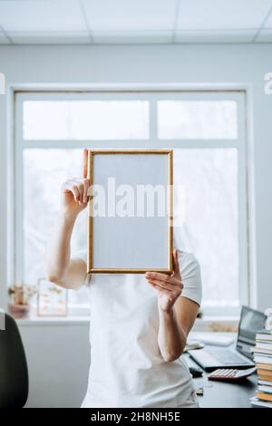 Frame picture, poster, diploma, certificate mockup in female hands. Faceless woman holding empty blank wooden photo frame on table with laptop and Stock Photo