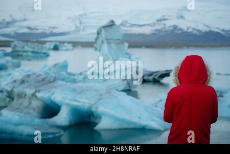 Rear view of female tourist, young woman in red jacket with a hood  looking at floating ice in Jokulsarlon glacier lagoon in Iceland, enjoying amazing Stock Photo
