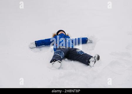 Best Outdoor Winter Activities for kids. Little kid boy in blue winter jacket making snow angel, laying down on snow in winter backyard. Active Stock Photo