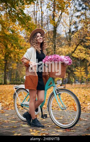 Pleasant bike ride along alleys of autumn park. Female beautiful bicyclist standing near retro street cycle with basket, filled by pink flowers at yellow town forest. Stock Photo