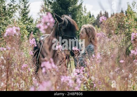 Young woman in meadow in flowers stroking brown horse. Stock Photo