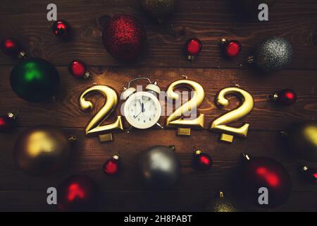 Candles in the form of numbers 2022, alarm clock showing midnight, multicolored christmas balls on a wooden background. Dark Christmas background. Stock Photo