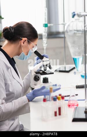 Scientist in goggles and medical mask using microscope near flasks and test tubes with blood samples in lab Stock Photo