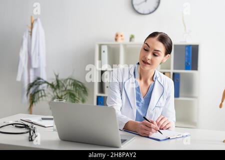 Doctor writing on clipboard and looking at laptop near eyeglasses and stethoscope in clinic Stock Photo