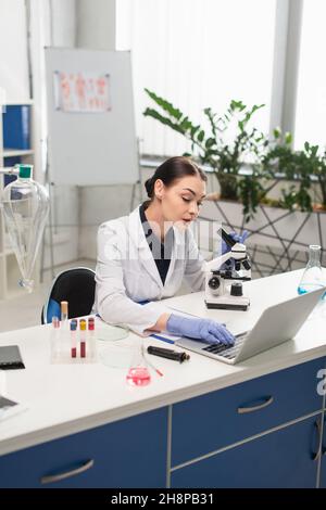 Brunette scientist using laptop near microscope and equipment in lab Stock Photo