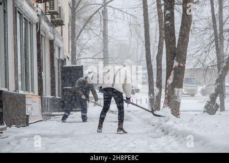 A girl dressed in light winter clothes is shoveling snow off the walkway. Behind her is a man with a shovel. Stock Photo