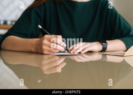 Close-up hands of unrecognizable young woman writing handwritten letter sitting at table at home, selective focus, blurred background. Stock Photo