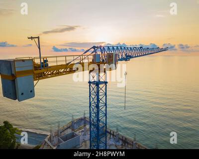 Close-up view from a drone of a construction tower crane against the background of a fantastic, beautiful sunset on the sea with a copy of the space Stock Photo