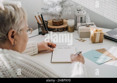 Wishlist, Modern Letters to Santa Claus from adults. Senior mature old woman in white sweater writing handwritten text Dear Santa in open notepad on Stock Photo