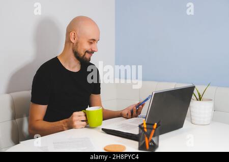Smiling bald man with beard in black T-shirt looking to phone in his hand and holding cap in another hand. Adult male working online on notebook at Stock Photo