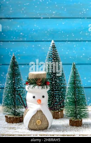 Christmas decoration snowman standing next to fir trees while it is snowing on a wooden table. Concept Christmas greeting card. Stock Photo