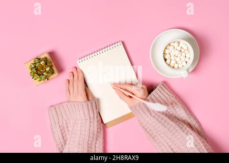 Notepad, golden tinsel, and a cup of hot chocolate with marshmallows and female's hands writing isolated on pink background. New year, Christmas or Stock Photo