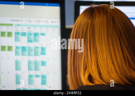 red haired female office worker in front of PC monitors with office calendar blurred on screen Stock Photo