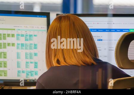 red haired female office worker in front of PC monitors with office calendar blurred on screen Stock Photo