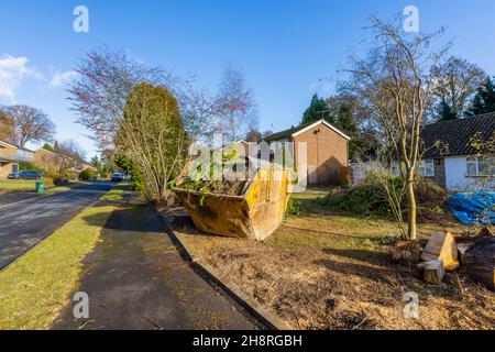 A large rusting, battered waste disposal skip full of branches and garden waste materials on the front garden of a road in Surrey, south-east England Stock Photo