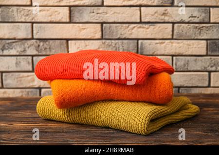 Stack of stylish sweaters on wooden table against brick wall background Stock Photo