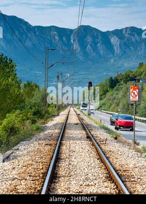 Perspective of the steel rails seemingly vanishing into mountains that surround Skadar Lake,in the morning sunlight of late summer,next to a busy high Stock Photo