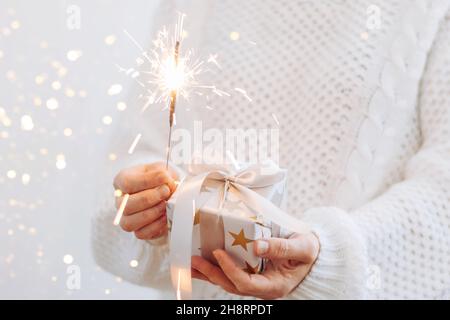 A girl in a white sweater holds a gift and sparkling sparklers. Holiday concept New Year or Christmas. Selective focus Stock Photo