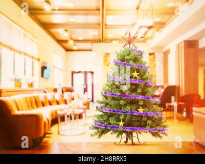 Artificial, Christmas tree with tinsel and stars stands on a blurred background of the hall with a large sofa. Bright photo, defocus. 3D rendering. Stock Photo