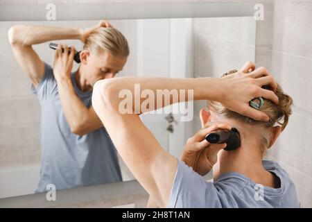 Man shaving whiskers hair by himself, self haircut at home in front of mirror in the bath. Male with long haircut using shaving machine trimmer Stock Photo