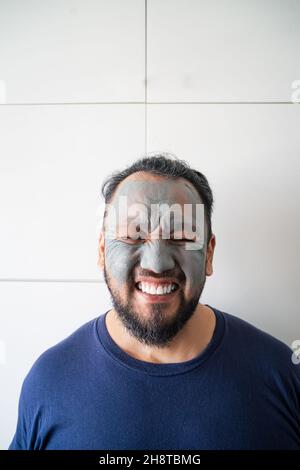 Beauty portrait of an excited matured Asian man standing isolated over white background, wearing facial cosmetic clay mask. Stock Photo