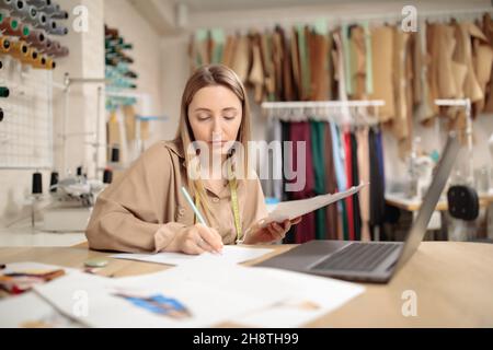 Got an idea. Interested millennial lady sitting at desk in workshop studio drawing sketch near laptop screen. Stock Photo