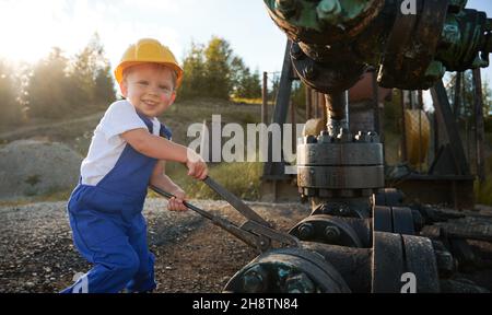Cute cheerful child looking at camera and smiling while tightening bolt on pipe in oil field. Joyful kid in construction helmet using industrial wrench while repairing pump jack pipe on sunny day. Stock Photo