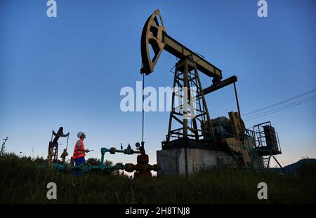 Male worker holding clipboard and looking at petroleum pump jack while working in oil field. Oil man standing near oil pumping unit under blue sky. Concept of oil extraction and petroleum industry. Stock Photo