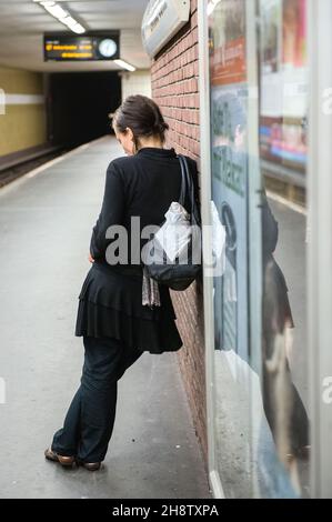 Berlin, Germany. Young adult woman leaning against an U-Bahn Station's interior wall while waiting for her connecting train towards Alexanderplatz. Meanwhile she is messaging connections using her Smartphone. Stock Photo
