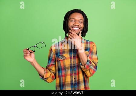 Photo of pretty thoughtful dark skin man wear plaid shirt smiling holding spectacles arm chin isolated green color background Stock Photo
