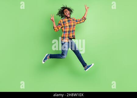 Photo of pretty funky dark skin man wear plaid shirt spectacles smiling jumping high showing v-signs isolated green color background