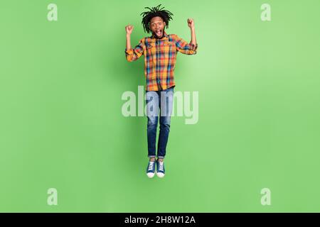 Photo of charming lucky dark skin man wear plaid shirt smiling jumping high rising fists isolated green color background