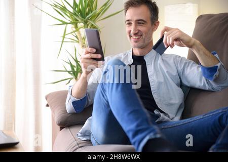 Happy man buying online from a smart phone lying on the sofa at home. Horizontal composition. Stock Photo