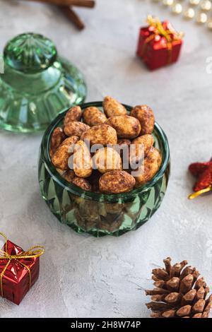 Candied or sugared almonds with honey and cinnamon in trendy green small glass jar on gray with Christmas decoration background. Traditional Christmas sweets for holidays. Copy space Stock Photo