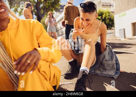 Female friends laughing together outdoors. Two young female friends having fun while sitting on the road in the summer sun. Group of young friends han Stock Photo