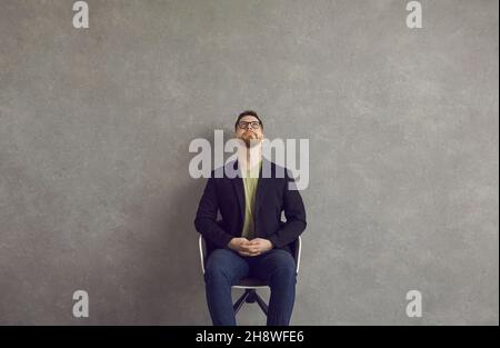 Young man sitting in chair near wall, looking up, thinking or imagining something Stock Photo