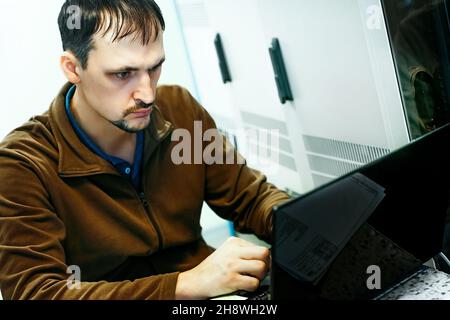 Portrait of a programmer at work. An authentic scene. An IT specialist has been working on a laptop in the server room for 30-40 years. Running diagnostics or performing maintenance work. Background. Stock Photo