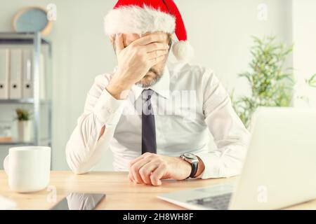 New Year depression, businessman in office wearing Santa Claus hat feeling lonely and depressed with hand covering face, selective focus Stock Photo