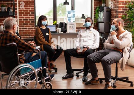 Multi ethnic people attending group therapy session during coronavirus pandemic. Old man sitting in wheelchair and patients in circle preparing for aa meeting discussion and counseling. Stock Photo