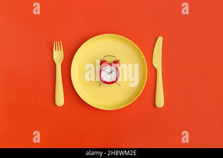 Red clock on the yellow plate with fork and spoon on a red background. Top view. Flat lay. Stock Photo