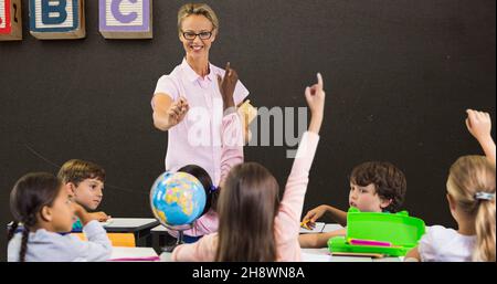 Smiling teacher pointing at students with hands raised while teaching in classroom Stock Photo