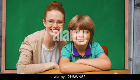 Portrait of smiling redhead teacher sitting with schoolboy against blackboard in classroom Stock Photo
