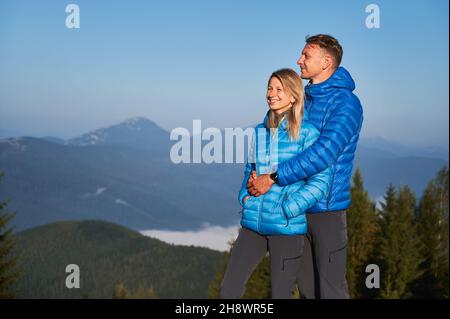 Handsome man hugging his smiling girlfriend from behind and they together standing and watching the surrounding nature against the mountain hills on sunny day. Stock Photo