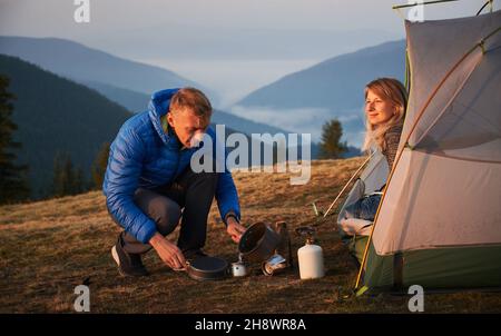 Camping in the morning. Hiker man sitting squatting and pouring water to make coffee near his wife, who admiring the surrounding beauty at dawn from their tent against misty mountains. Stock Photo
