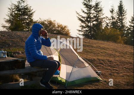 Side view of boy in hooded jacket drinking coffee on wooden bench in his camp near tent set up on mountain hill meadow. Autumn hiking and camping in nature. Concept of hiking, travelling and camping. Stock Photo