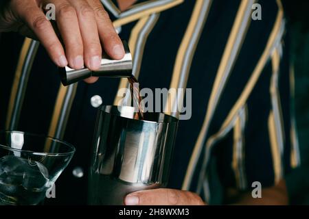 a young caucasian man, wearing a striped shirt, is about to prepare a cocktail in a metal shaker by adding a dark liquor to it with a jigger Stock Photo